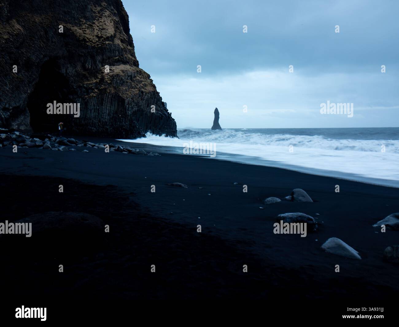 Spiaggia di Reynisfjara al crepuscolo, caratterizzata da una spettacolare sabbia nera, grotta di basalto, scogliere aspre e le iconiche faraglioni di Reynisdrangar che emergono dalla turbul Foto Stock