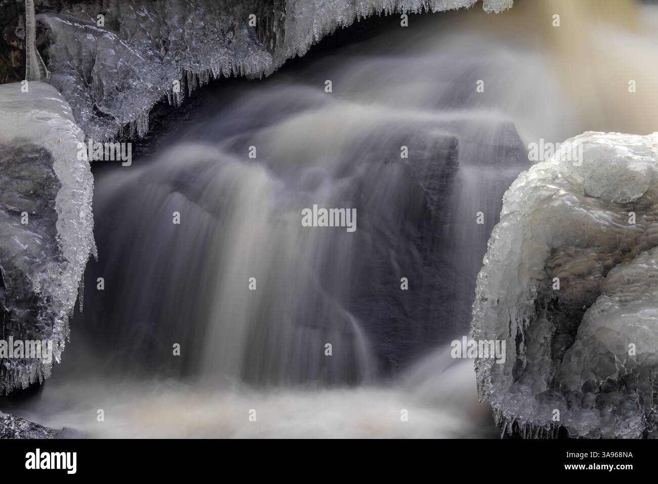 Primo piano di una cascata ghiacciata con acqua corrente nebbiosa, piccoli ghiaccioli raggruppati appesi alle rocce e formazioni di ghiaccio che coprono la superficie di pietra Foto Stock