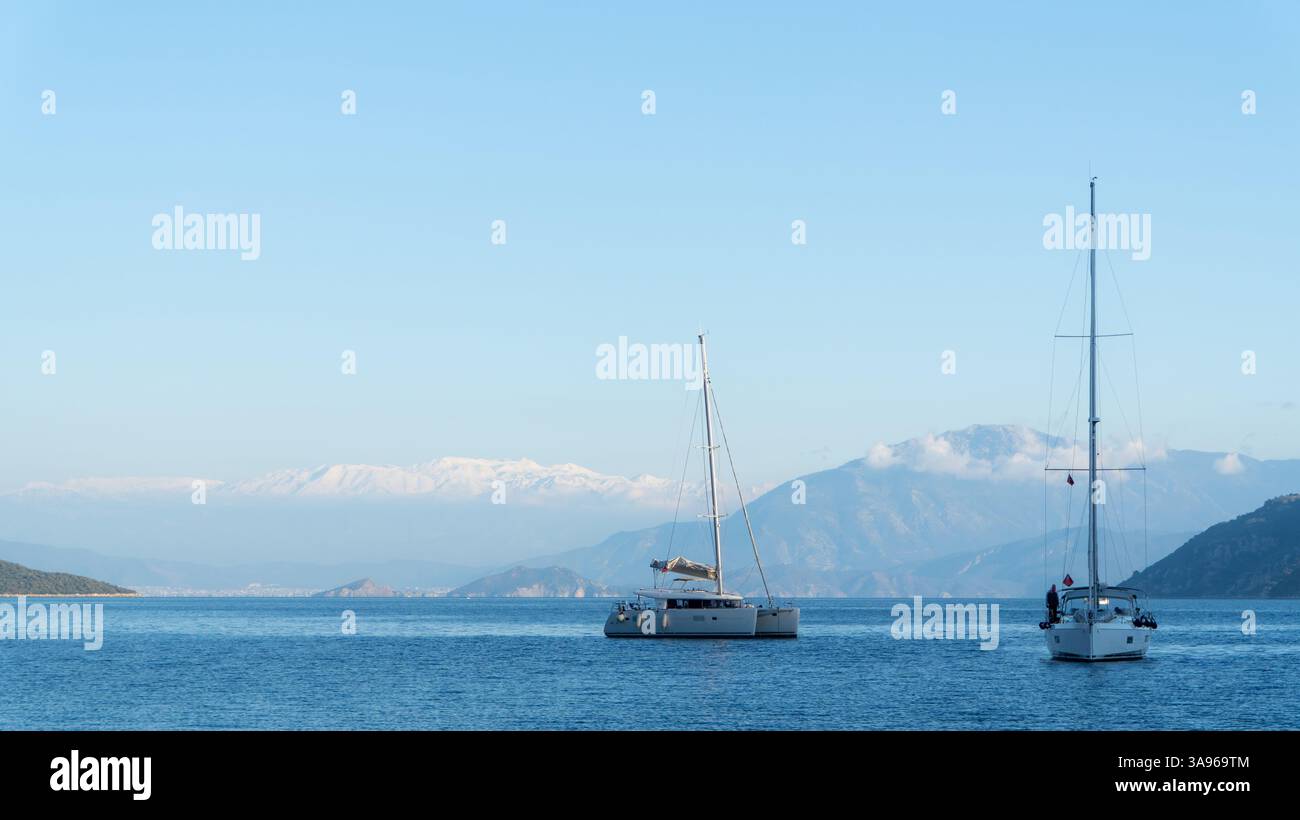 Due barche a vela navigano sul Mar Mediterraneo a Fethiye, in Turchia, con uno splendido sfondo di montagne innevate e un cielo azzurro limpido, creando un'atmosfera unica Foto Stock