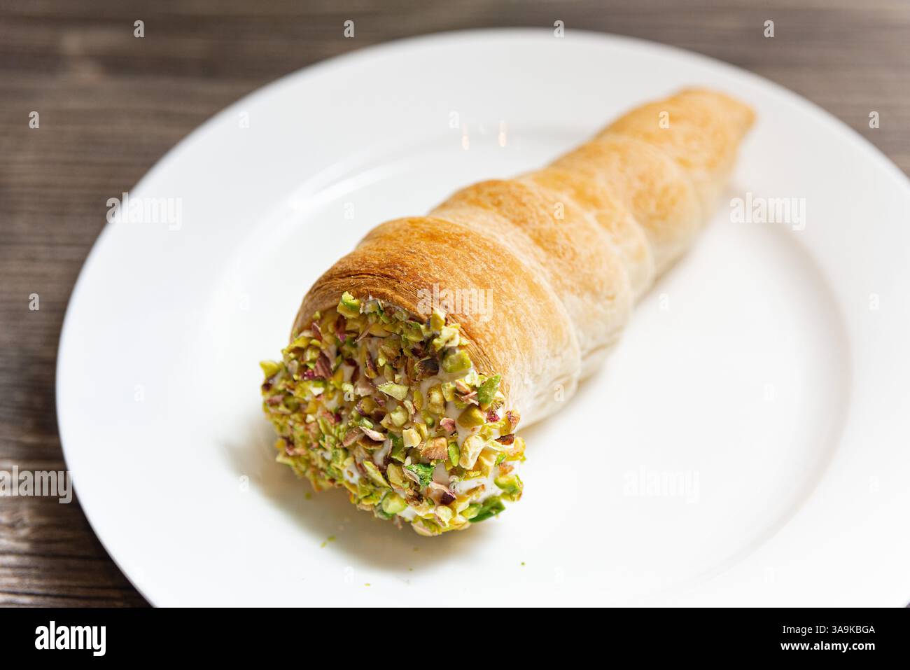 Un corno di pasticceria dorata, pieno di panna e ricoperto di pistacchi schiacciati, servito su un piatto bianco su un rustico tavolo di legno Foto Stock