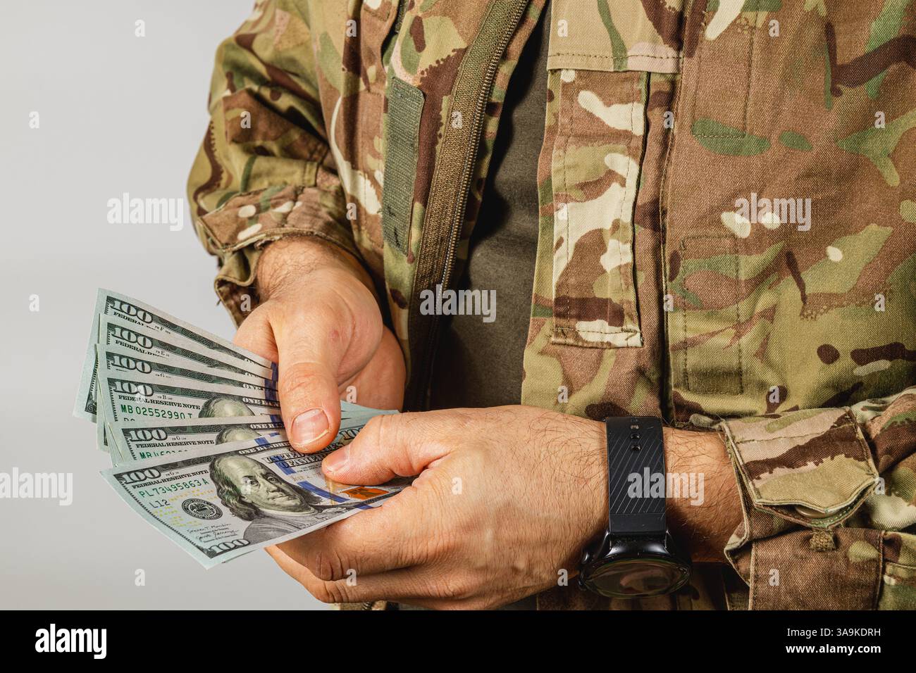 Un soldato in abiti mimetici conta più banconote mentre si trova in un ambiente neutrale, illustrando un momento focalizzato sulla gestione del contante. Foto Stock