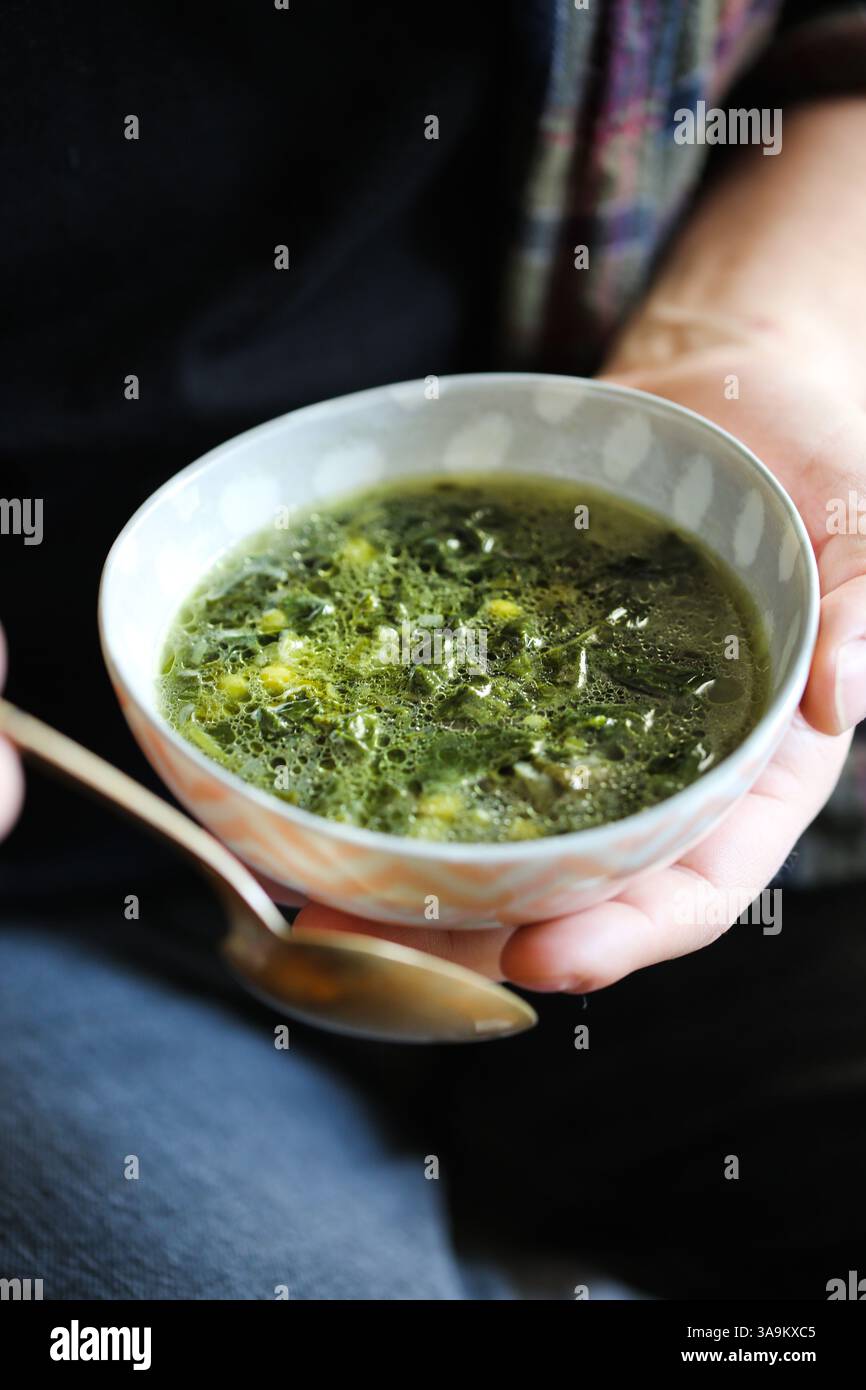 Ciorbă di spinaci primaverili rumena servita in una ciotola e tenuta tra le mani. Zuppa tradizionale dell'Europa orientale a base di spinaci novelli, cipollotti e aneto. Foto Stock