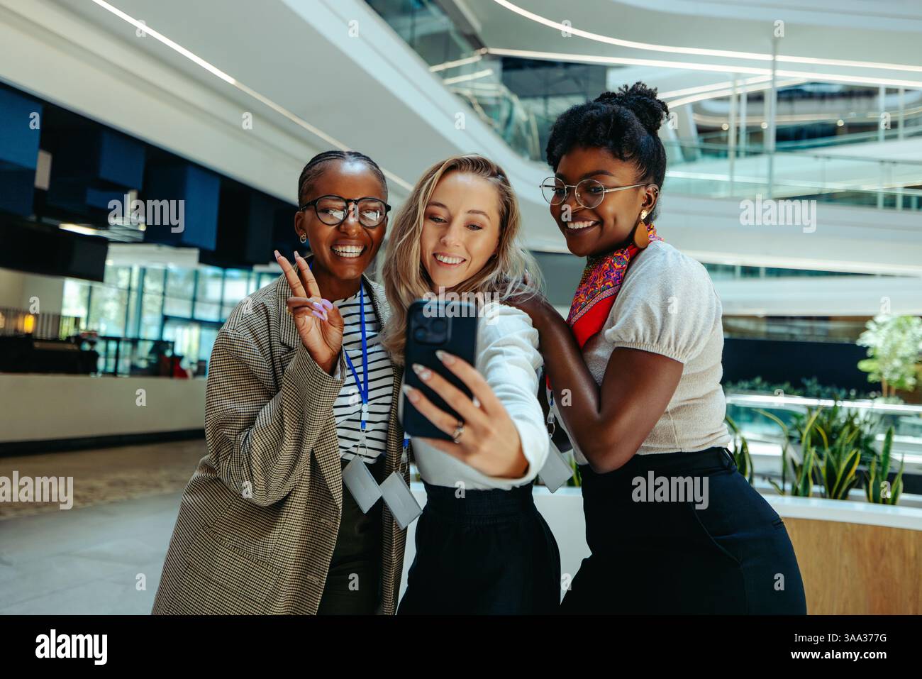 Giovani reclute eccitate si mettono in posa per un selfie in un ufficio elegante, celebrando una nuova opportunità di lavoro con gioia e lavoro di squadra. Foto Stock