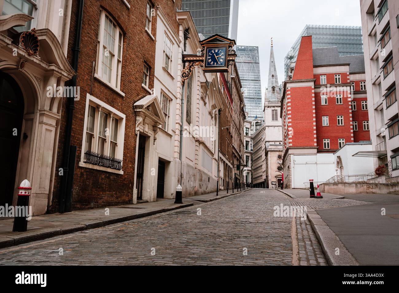 Una tranquilla strada londinese con ciottoli che si snoda verso una chiesa e torri di vetro, con uno storico orologio sospeso Foto Stock