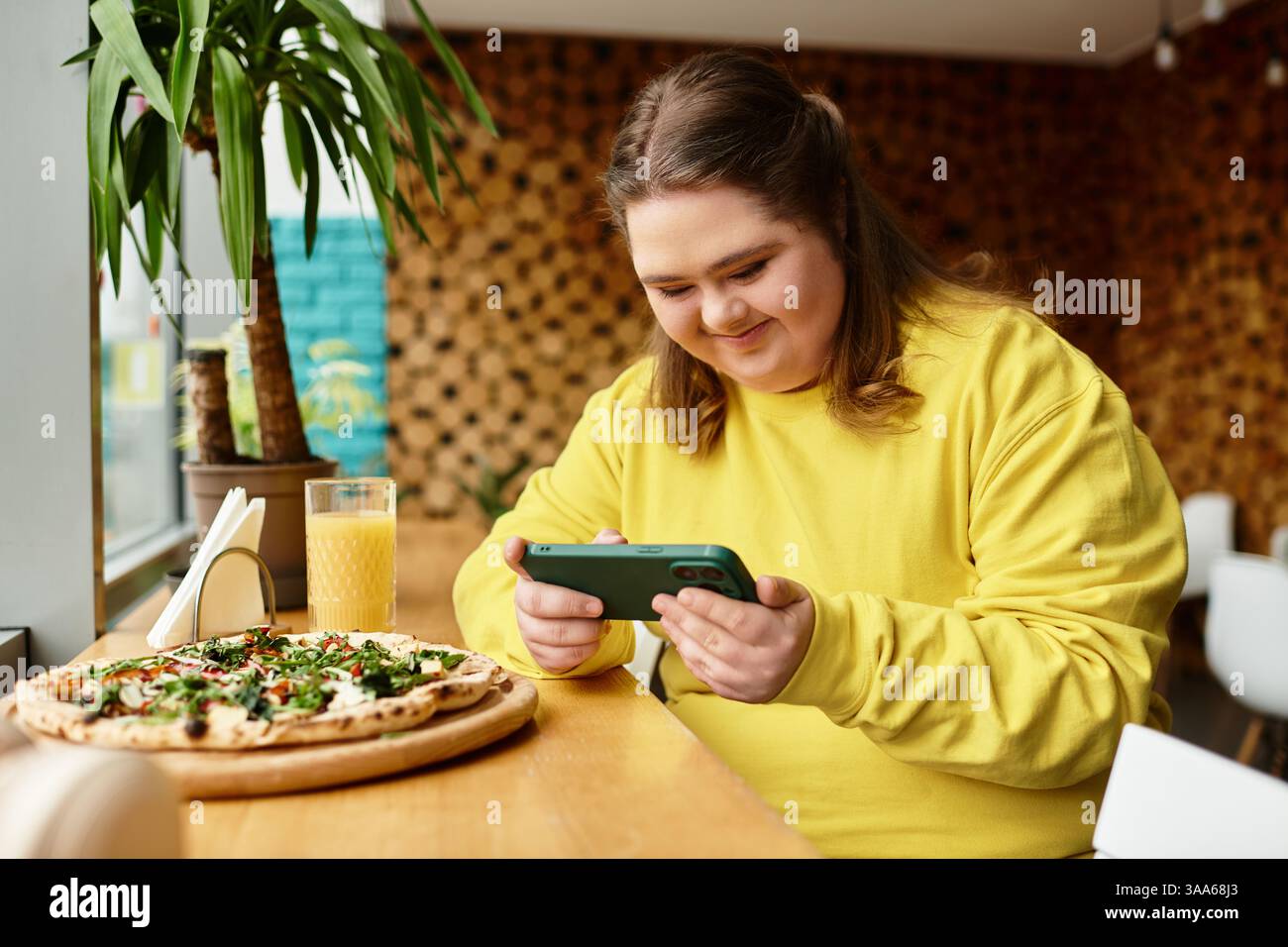 Una giovane donna con la sindrome di Down sorride al telefono mentre assapora un pasto delizioso in un bar. Foto Stock