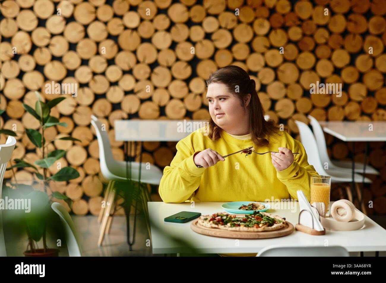 La giovane donna affetta da sindrome di Down siede con fiducia a un tavolo, gustando il suo pasto e bevendo. Foto Stock