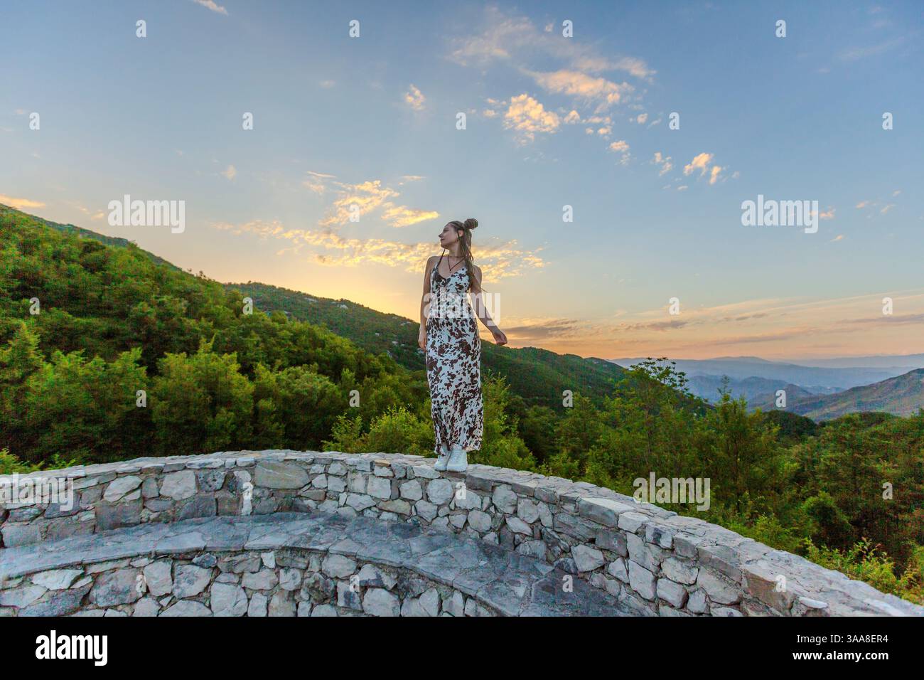 Una donna con un abito che scorre si erge su una piattaforma di pietra, affacciata su un vasto paesaggio al tramonto. La sua silhouette contrasta con il cielo colorato Foto Stock