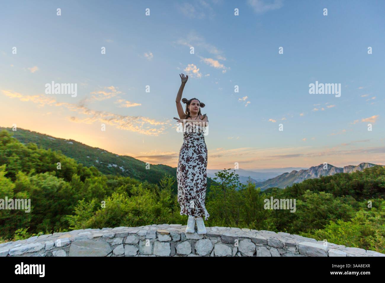 Una figura in un abito che scorre si erge su una struttura in pietra, sollevando un braccio verso un vibrante tramonto. L'immagine mostra una miscela di grazia umana Foto Stock