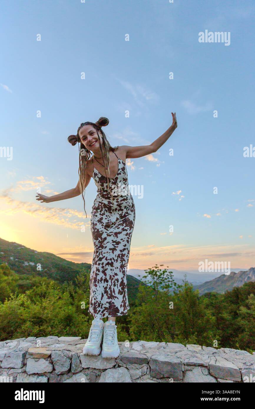 Una donna in un abito che scorre si erge in cima ad una struttura di pietra al tramonto, braccia sollevate con grazia. La sua silhouette contrasta con il cielo vibrante Foto Stock