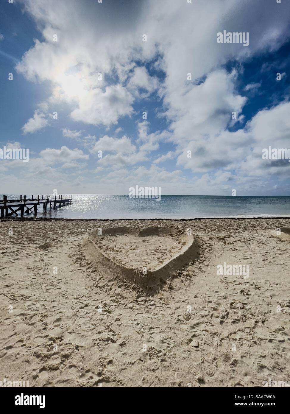 Un design a forma di cuore scolpito nella sabbia su una spiaggia tropicale soleggiata, con un molo e un oceano sullo sfondo. Perfetto per viaggi e temi romantici. - Immagine stock catturata con smartphone
