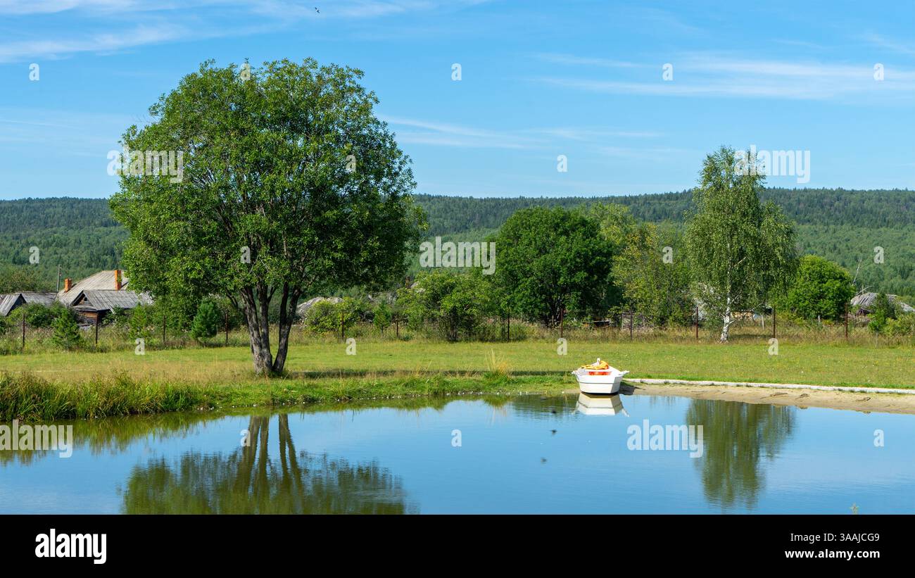 Piccola barca bianca da diporto non motorizzata o senza motore sulla riva del laghetto o del lago nel villaggio o nell'area rurale. Paesaggio rurale pastorale. Attività ricreative all'aperto Foto Stock