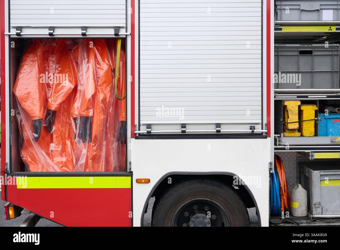 Primo piano di un compartimento per camion antincendio contenente tute hazmat e attrezzature di risposta alle emergenze. Le tute protettive di colore arancione brillante sono sigillate in plasti Foto Stock