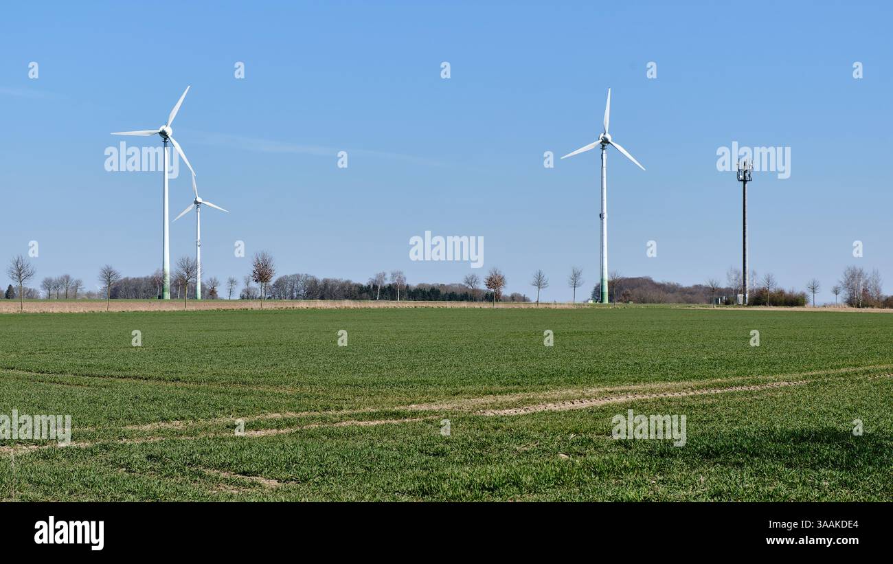 Tre vecchie turbine eoliche e un albero di telecomunicazioni si trovano in un paesaggio agricolo verde nella bassa Sassonia, in Germania, sotto un cielo blu brillante, SYM Foto Stock