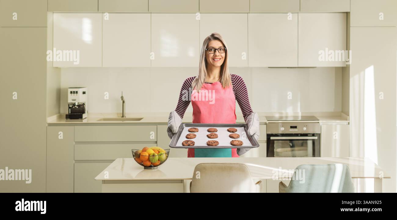 Giovane donna con un grembiule che tiene un vassoio di biscotti appena sfornati in una cucina Foto Stock