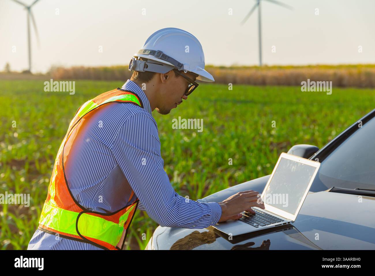 Giovane ingegnere professionista che lavora su un computer portatile di fronte al cofano auto in campagna all'aperto con sfondo di un parco di turbine eoliche Foto Stock Giovane ingegnere professionista che lavora su un computer portatile di fronte al cofano auto in campagna all'aperto con sfondo di un parco di turbine eoliche Foto Stock