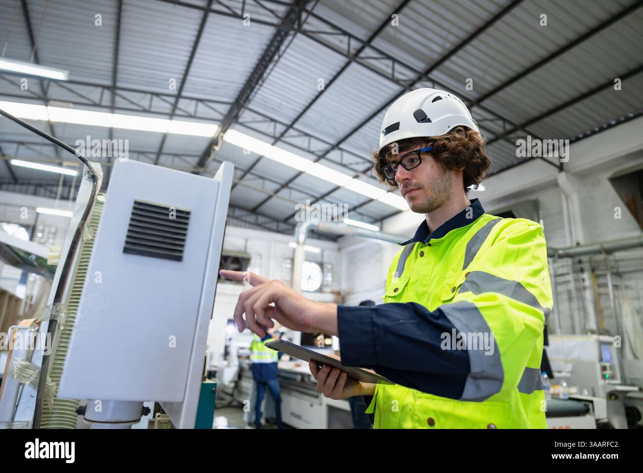 Il giovane controllo del lavoratore maschile caucasico aziona la macchina CNC della taglierina del legno nell'industria della fabbrica di mobili con tuta di sicurezza Foto Stock