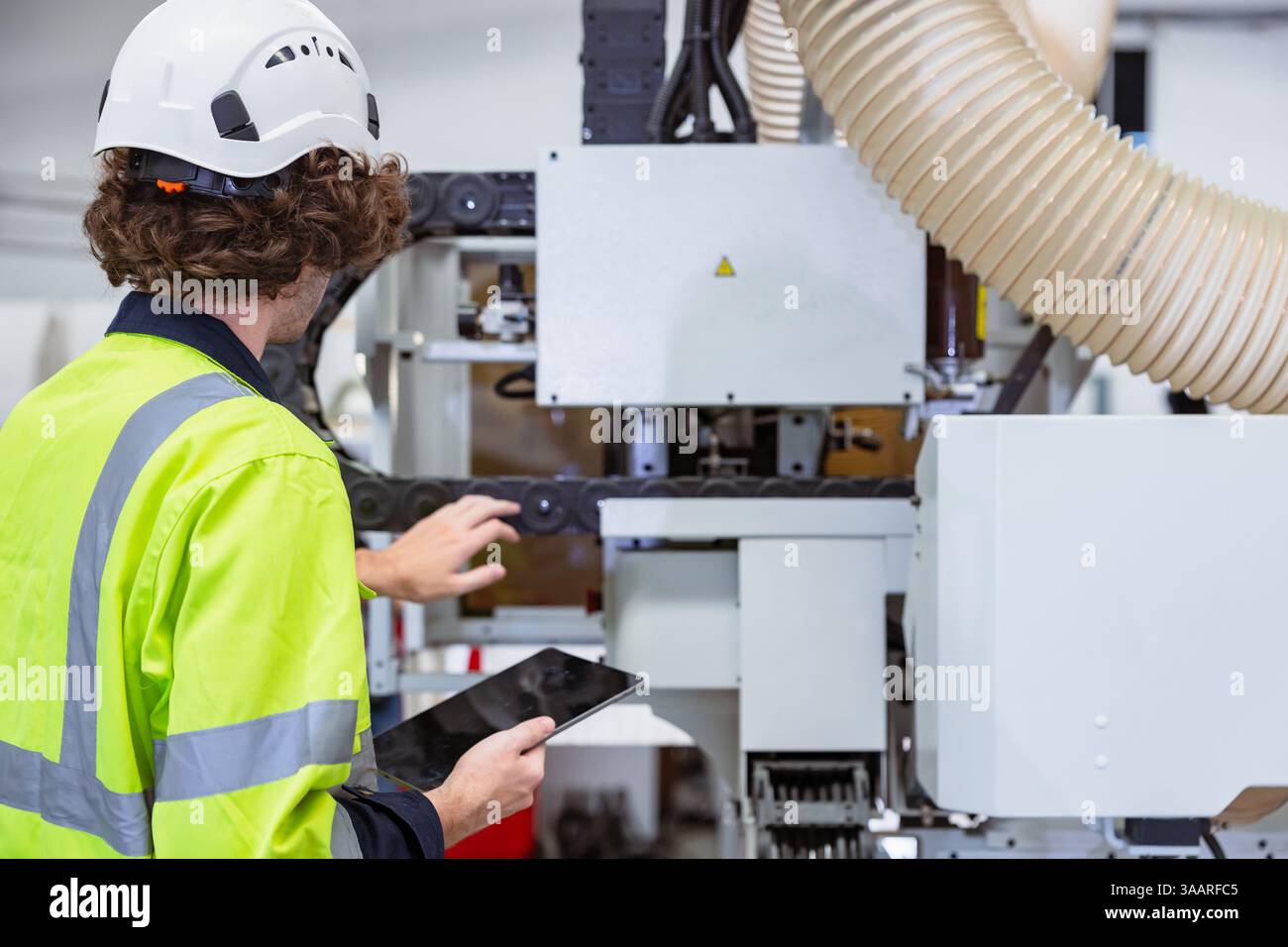 Il giovane controllo del lavoratore maschile caucasico aziona la macchina CNC della taglierina del legno nell'industria della fabbrica di mobili con tuta di sicurezza Foto Stock