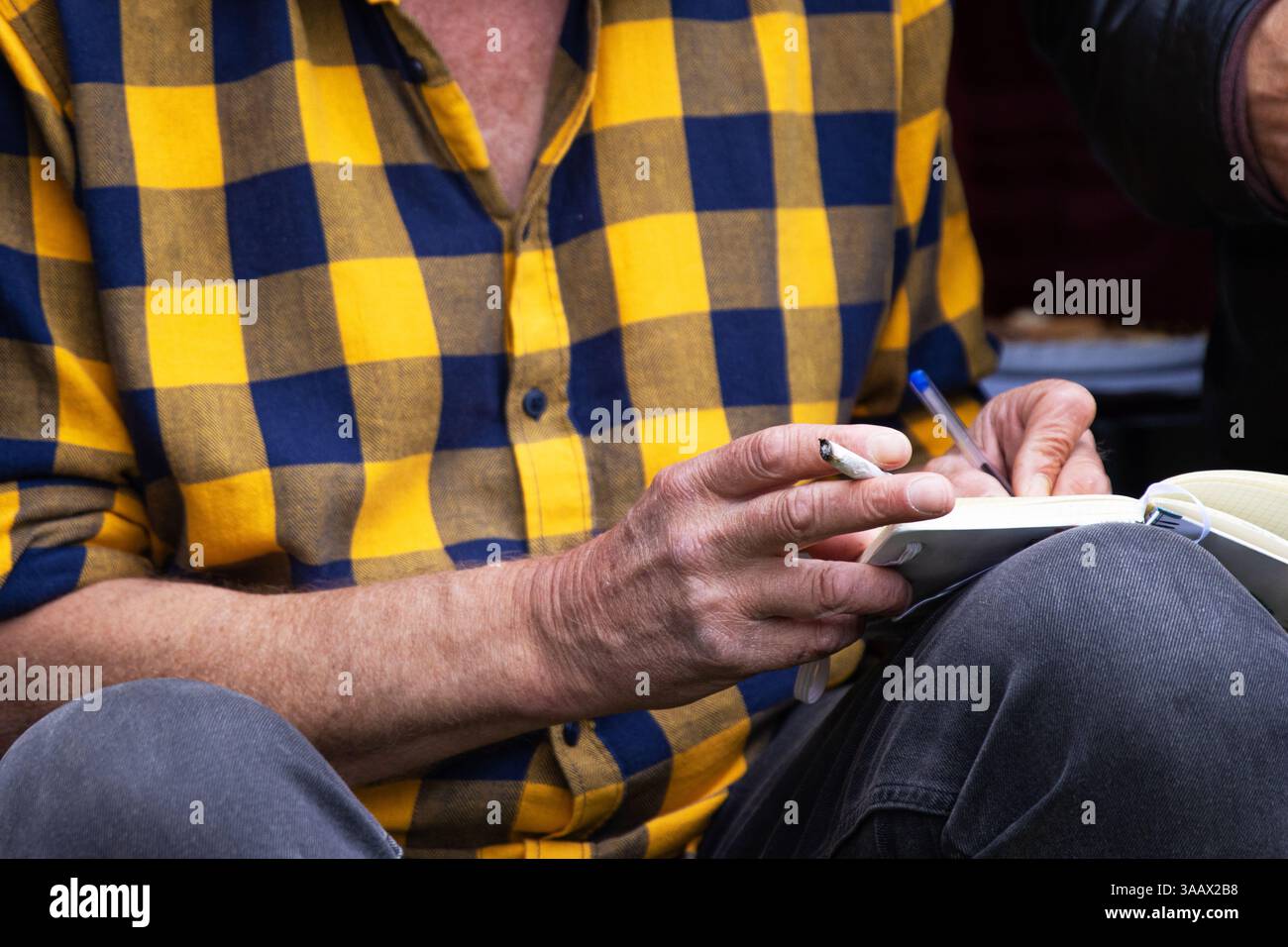 Primo piano di un uomo con una camicia gialla a scacchi che tiene una penna e una sigaretta, che scrive su un taccuino abitudini di fumo, creatività, prendere appunti, scrivere diari, Foto Stock
