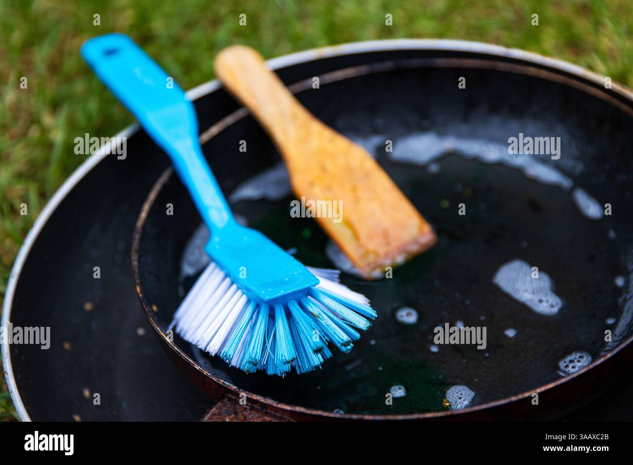 Utensili da cucina per il risciacquo all'aperto. Padella sporca con schiuma di sapone, una spatola di legno e un pennello blu sull'erba verde. Lo era lo stile di vita minimalista del campeggio Foto Stock