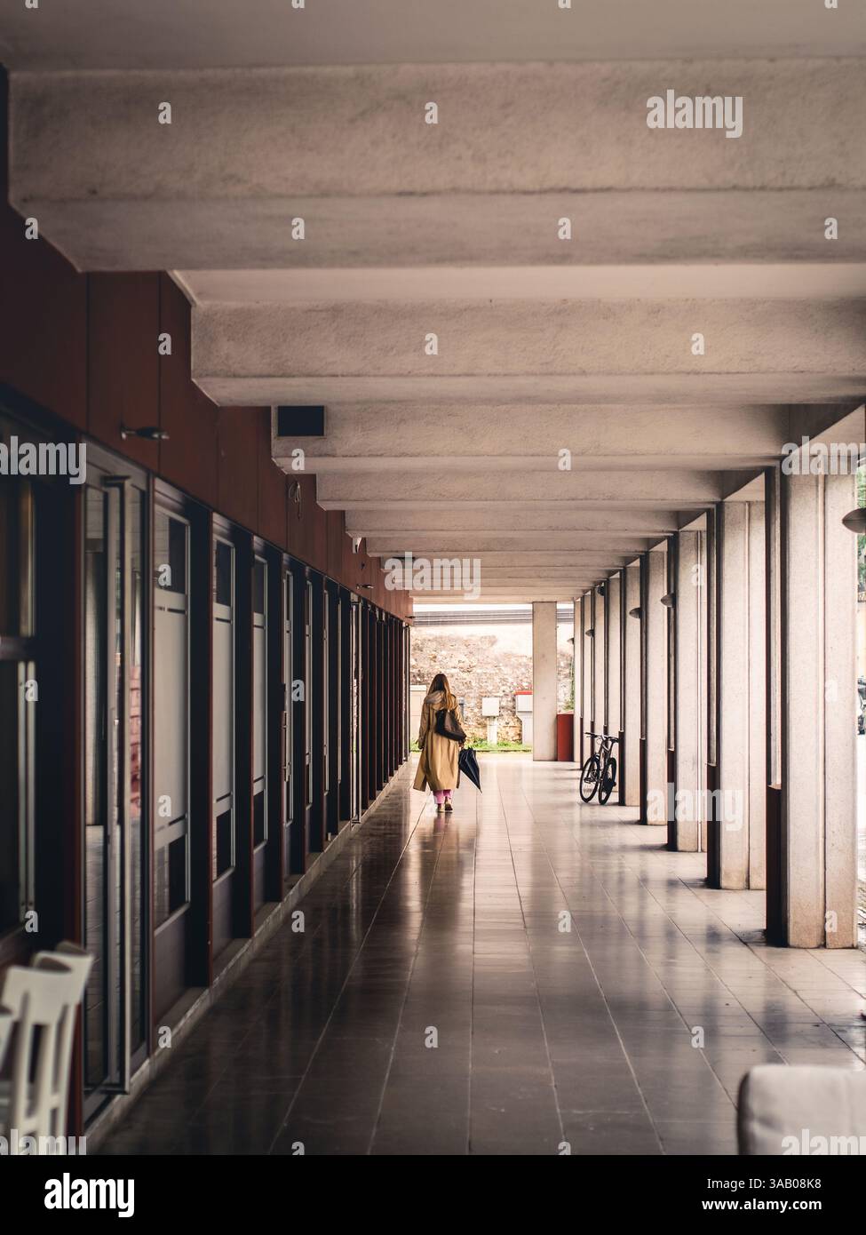 Una persona con un cappotto beige cammina attraverso una moderna passerella coperta in Italia. Il soffitto geometrico e le colonne creano profondità, mentre una bicicletta si inclina Foto Stock