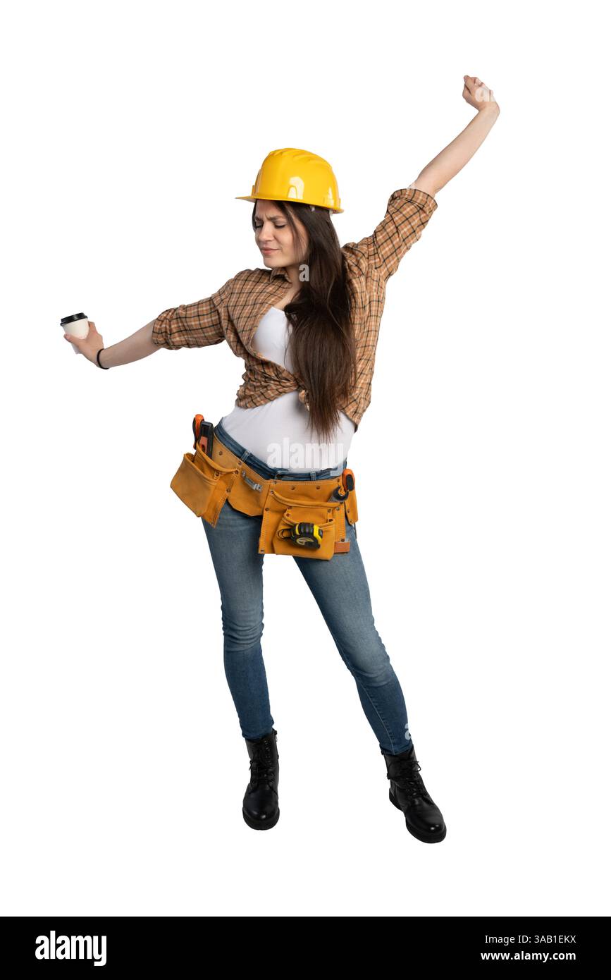 Lavoratrice edile che indossa un elmetto giallo e una cintura portautensili, che si allunga mentre tiene in mano una tazza di caffè, sembra stanca ma pronta per il lavoro. Foto Stock