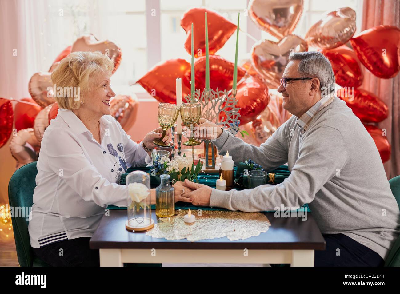 Una romantica coppia anziana che festeggia San Valentino o anniversario a casa, brindando con i bicchieri e tenendo per mano un tavolo decorato con il cuore Foto Stock