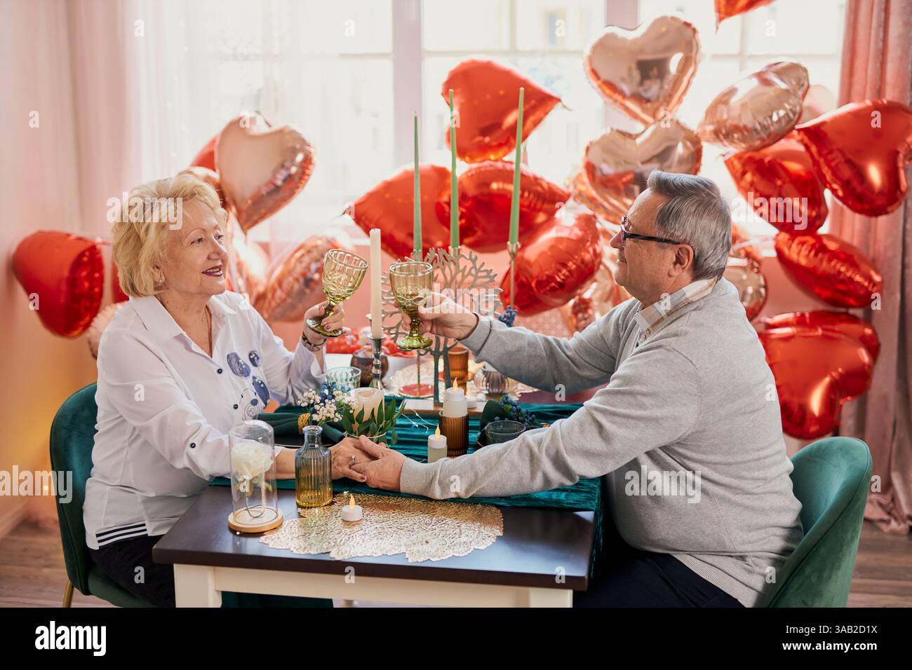 Una romantica coppia anziana che festeggia San Valentino o anniversario a casa, brindando con i bicchieri e tenendo per mano un tavolo decorato con il cuore Foto Stock