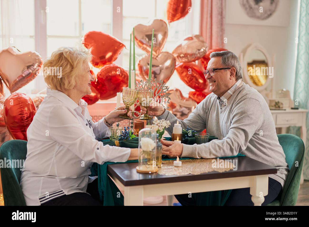 Una romantica coppia anziana che festeggia San Valentino o anniversario a casa, brindando con i bicchieri e tenendo per mano un tavolo decorato con il cuore Foto Stock