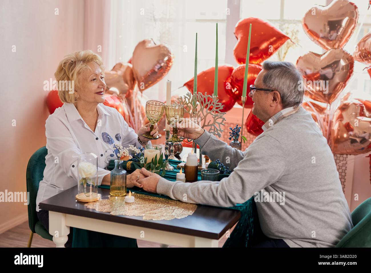 Una romantica coppia anziana che festeggia San Valentino o anniversario a casa, brindando con i bicchieri e tenendo per mano un tavolo decorato con il cuore Foto Stock