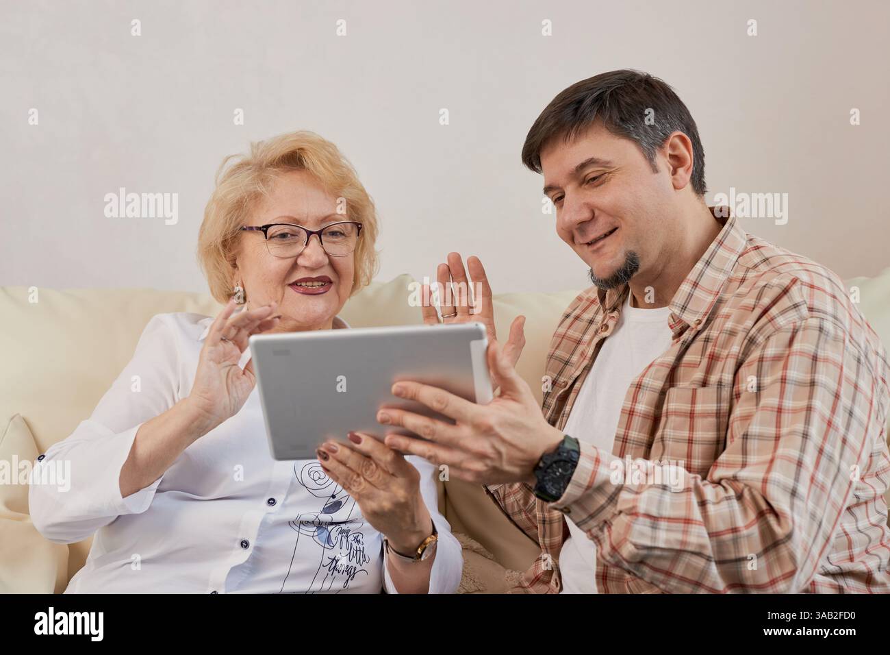 Giovane che aiuta una madre anziana a usare un tablet su un divano a casa. Connessione intergenerazionale, formazione tecnologica e concetto di famiglia Foto Stock