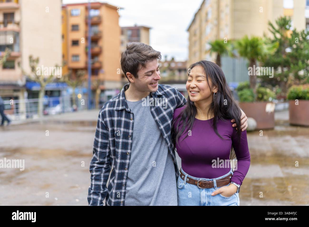 Una coppia multietnica cammina braccio nel braccio, condividendo un momento di gioia in un ambiente urbano. La scena cattura un mix di culture e felicità Foto Stock