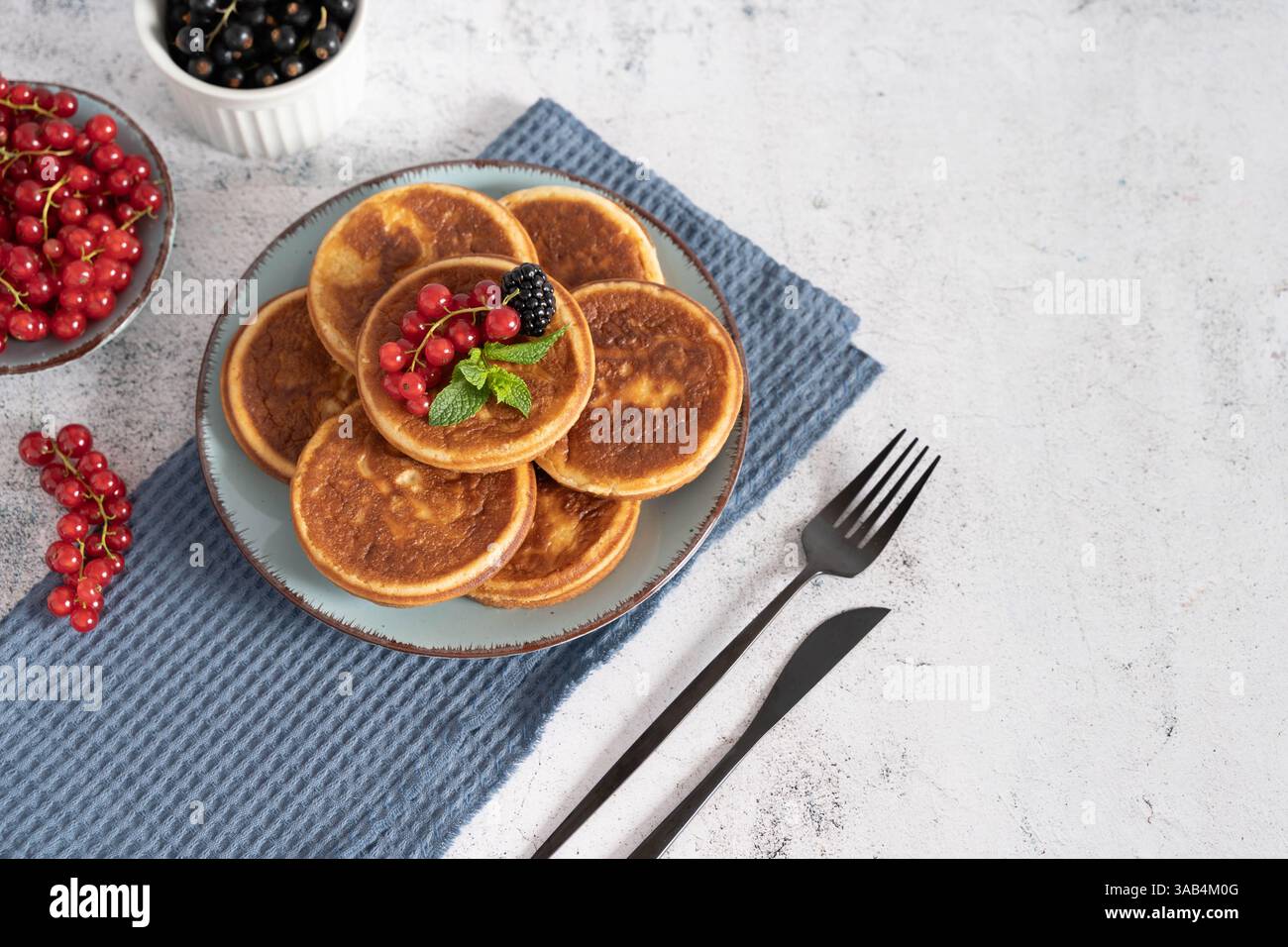 Pancake fatti in casa con mirtilli, lamponi e fragole su un piatto bianco con un panno blu su un tavolo grigio chiaro Foto Stock