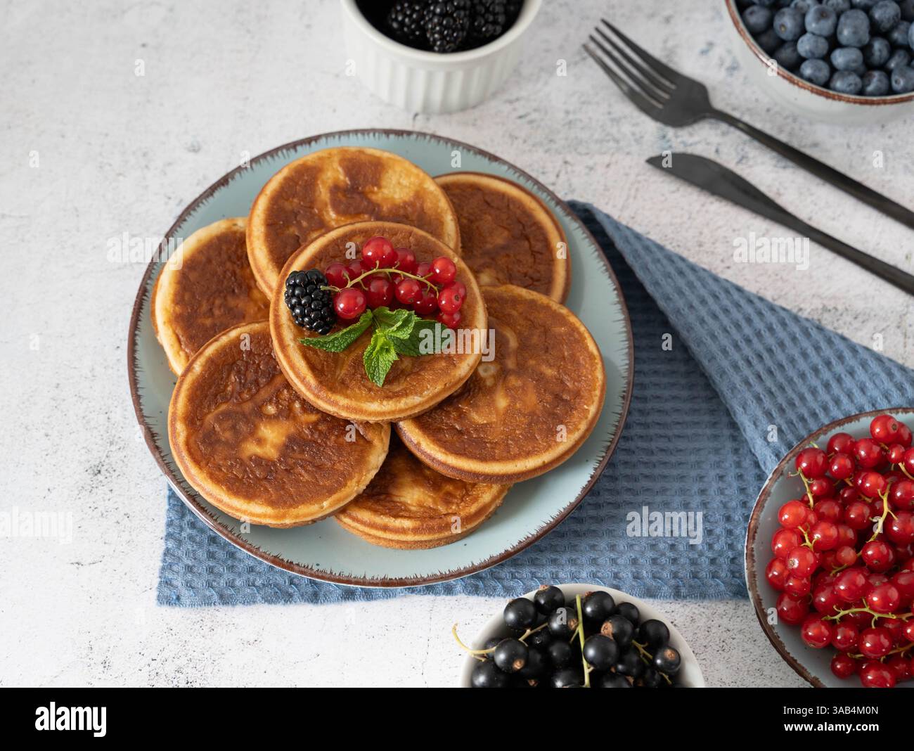 Pancake fatti in casa con mirtilli, lamponi e fragole su un piatto bianco con un panno blu su un tavolo grigio chiaro Foto Stock