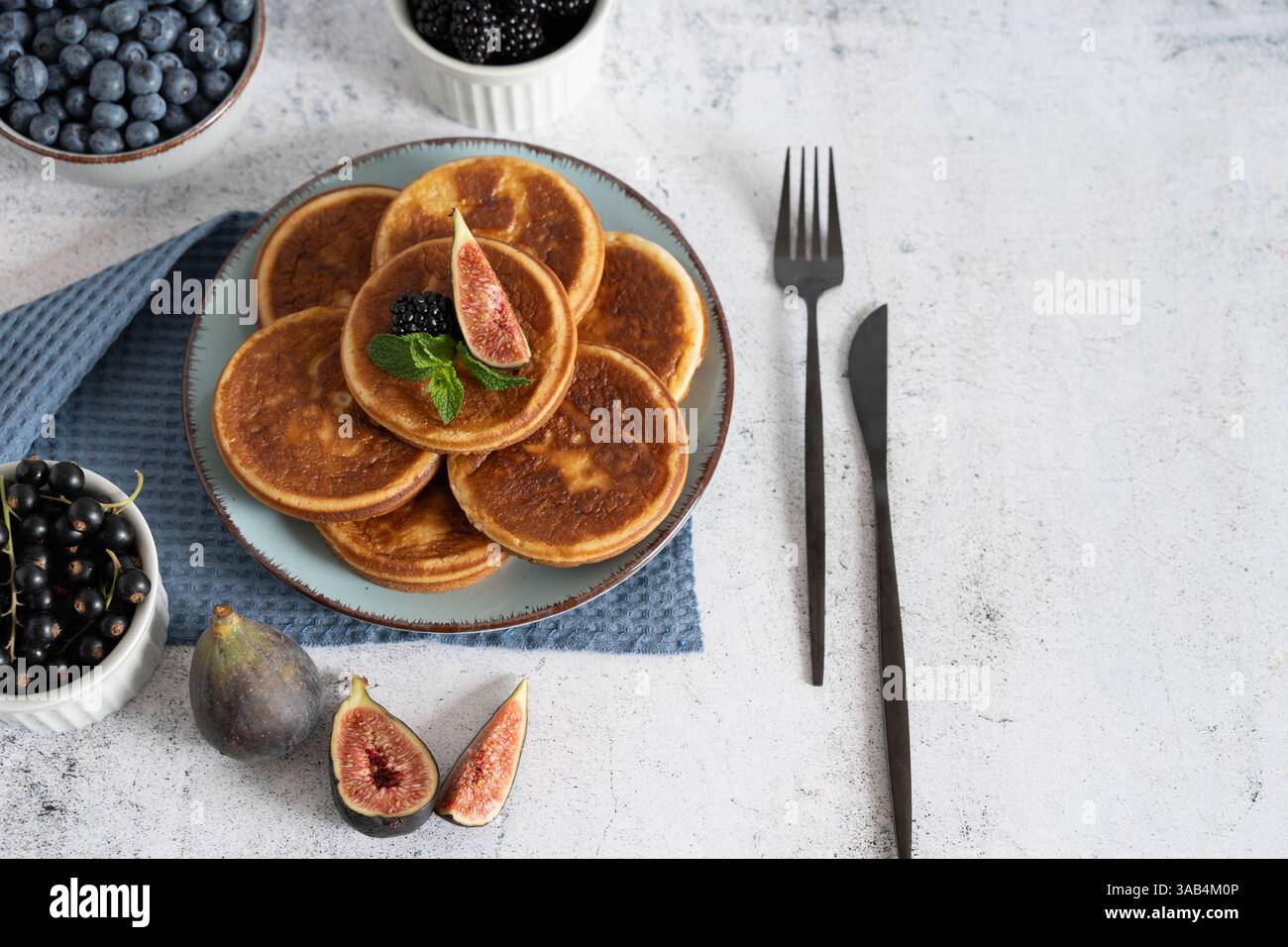 Pancake fatti in casa con mirtilli, lamponi e fragole su un piatto bianco con un panno blu su un tavolo grigio chiaro Foto Stock