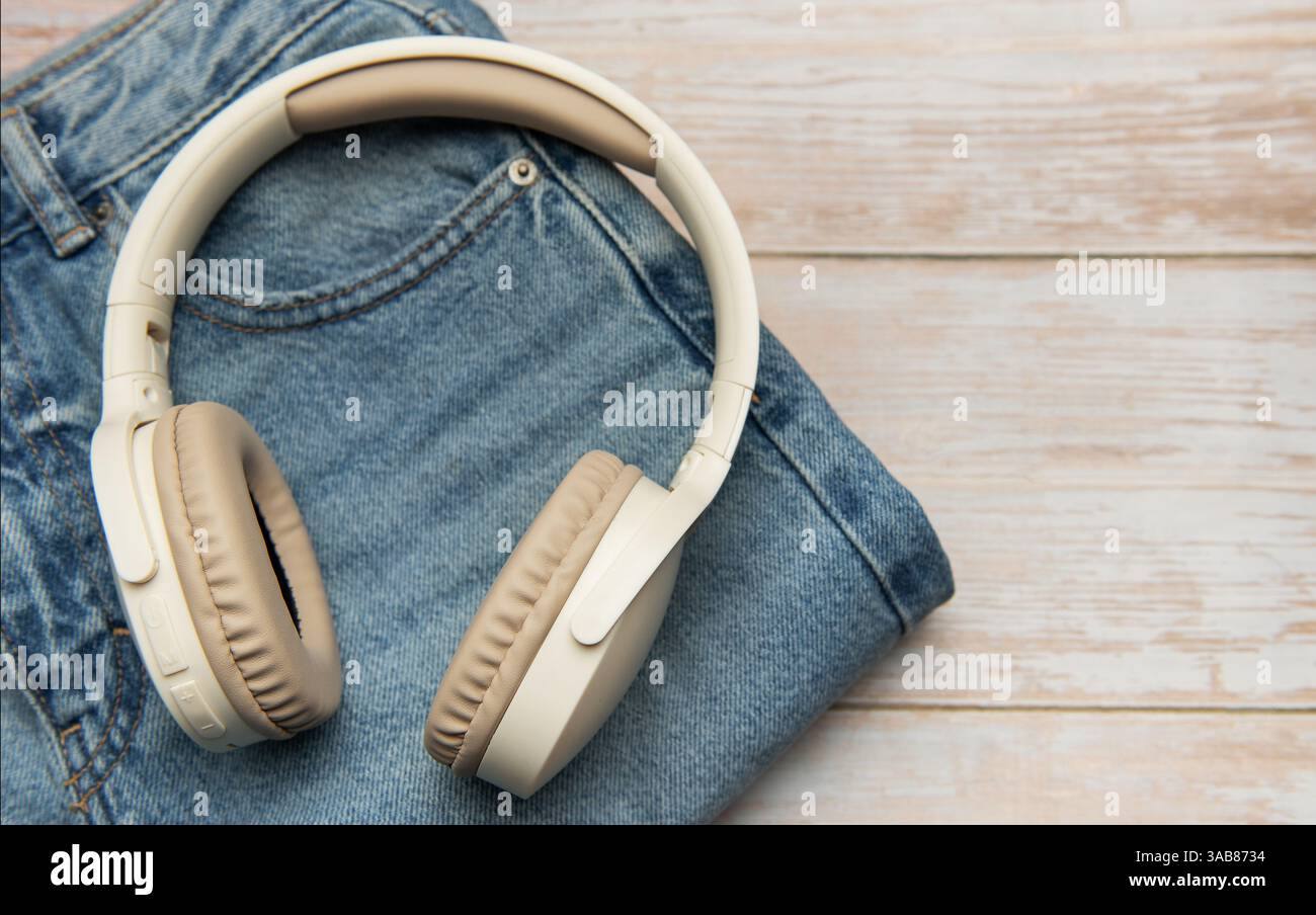 Moderne cuffie wireless posizionate su jeans in denim piegati su uno sfondo di legno chiaro, per creare un'immagine elegante e alla moda Foto Stock