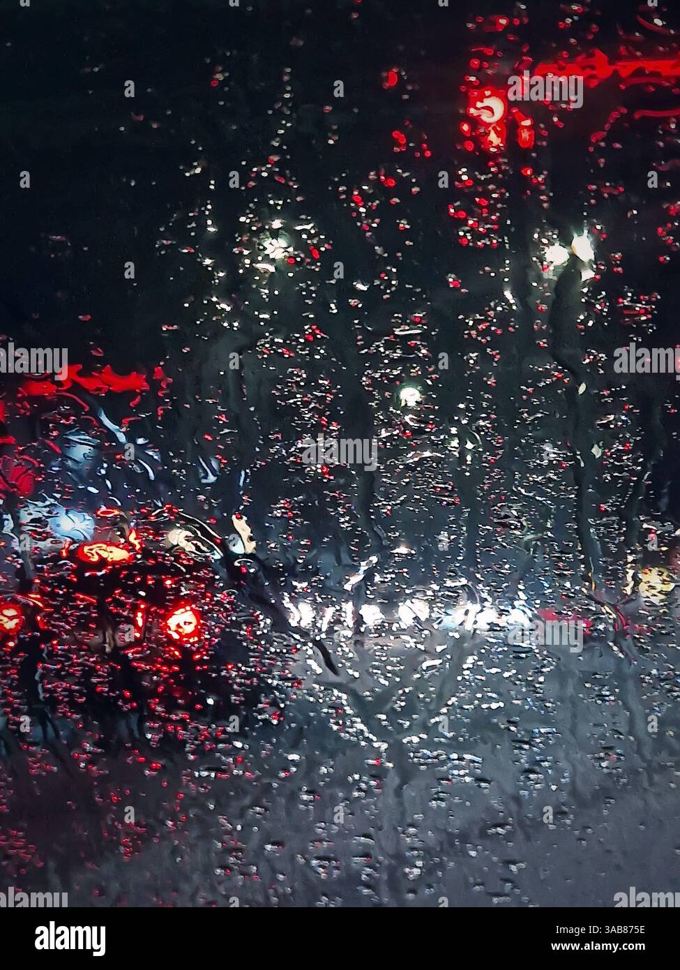 La scena del traffico urbano notturno d'inverno è visibile attraverso il parabrezza del veicolo coperto da gocce di nevischio. Lo scioglimento dell'acqua di neve cade sul finestrino dell'auto - Immagine stock catturata con smartphone