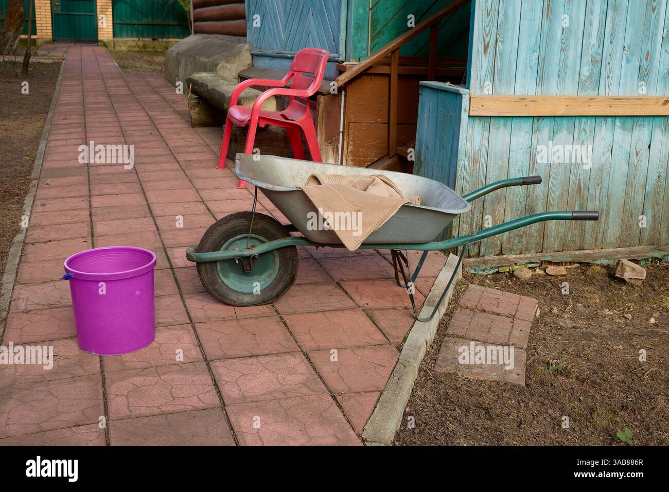 Carriola con coperta beige e secchio rosa su un sentiero pavimentato vicino a una casa di legno. Una sedia di plastica rossa si siede lateralmente, aggiungendo contrasto alla r Foto Stock