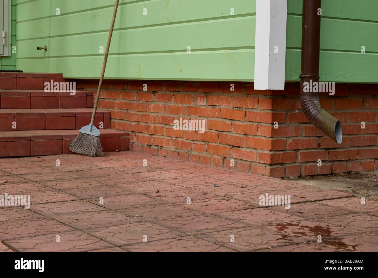 Una scopa si appoggia su gradini di mattoni rossi e su un muro, suggerendo un'operazione di pulizia all'aperto completata. L'ambientazione include un rivestimento in legno verde e una visiera Foto Stock