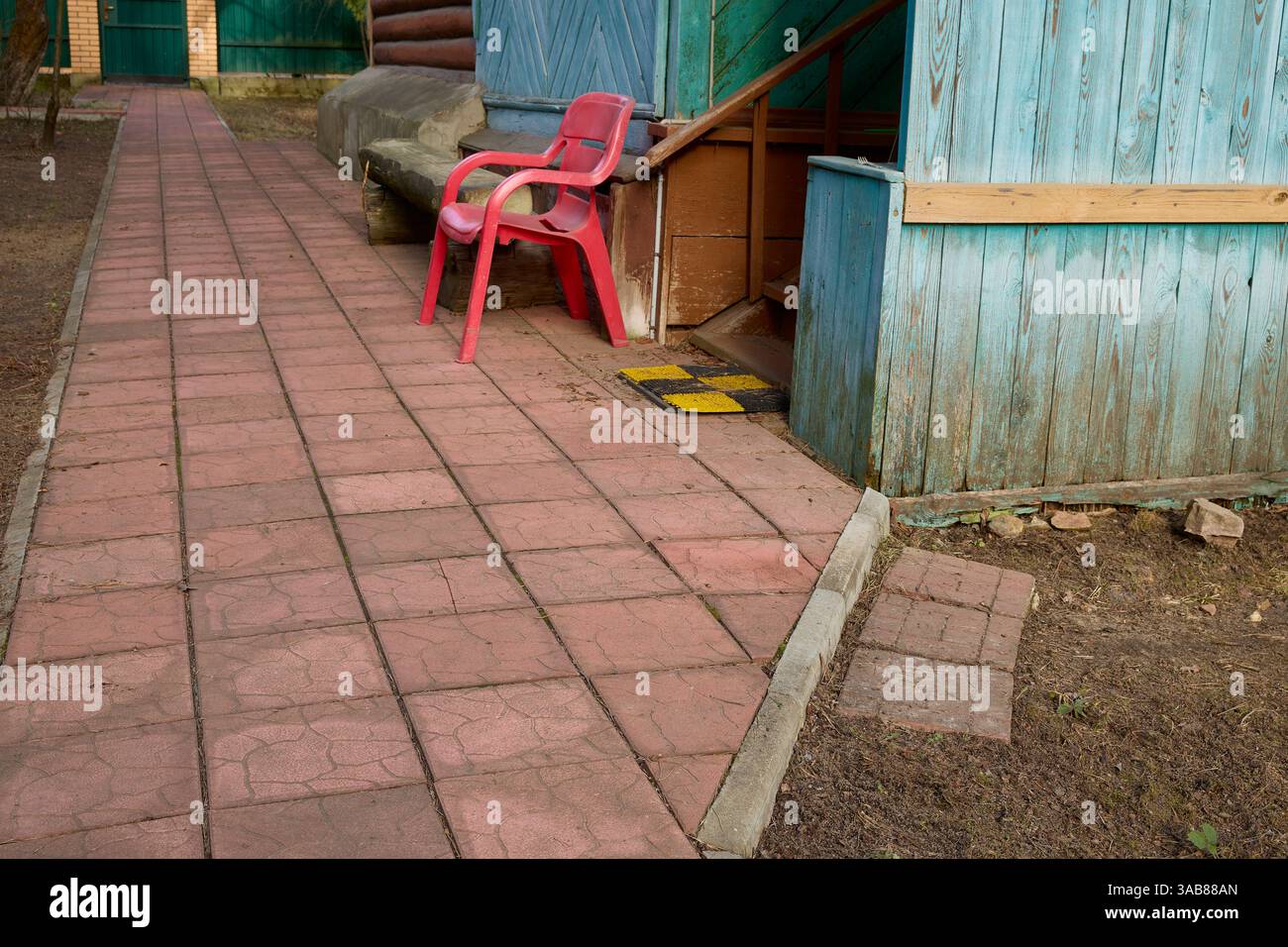 La sedia di plastica rossa poggia accanto a un edificio di legno blu vicino a un passaggio pedonale pavimentato. La scena all'aperto suggerisce un ambiente tranquillo e tranquillo, mettendo in risalto il carattere rustico Foto Stock