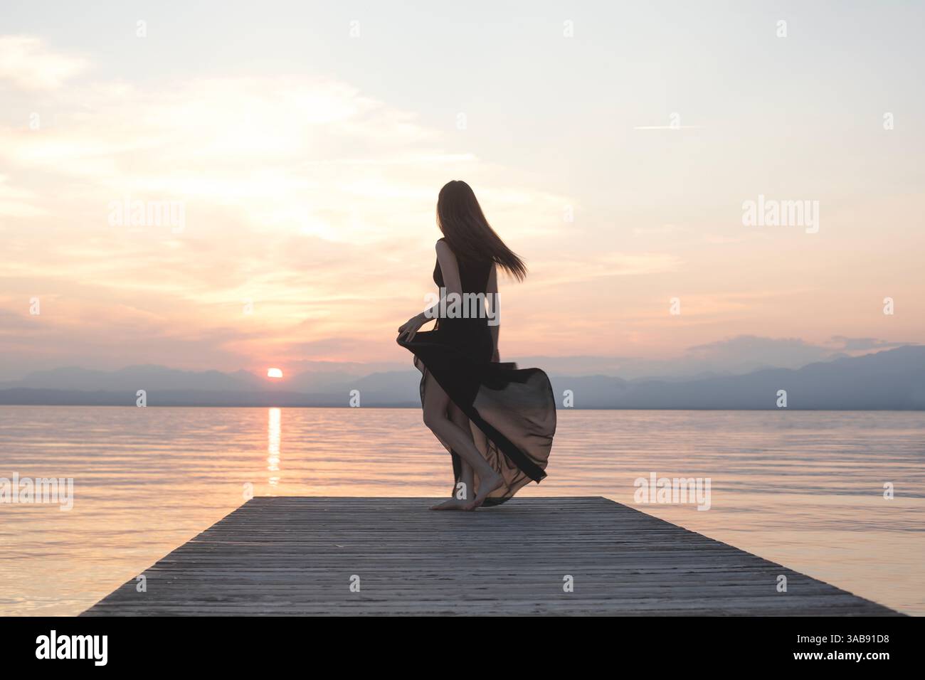 donna con un lungo abito nero che balla felicemente su un molo di fronte al mare al tramonto Foto Stock