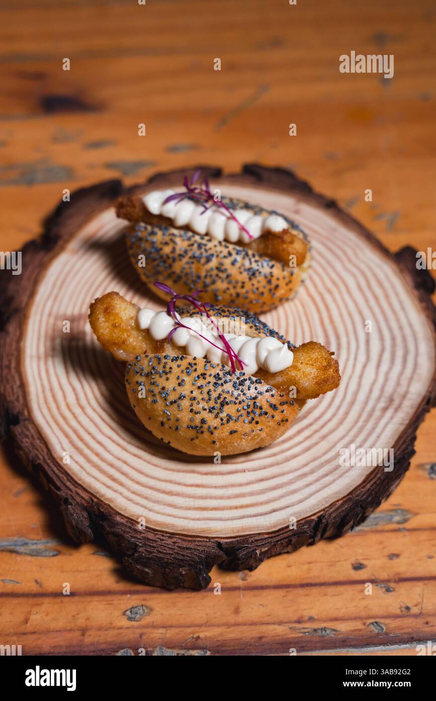 Due mini panini gourmet con semi di papavero, ripieni di fritti croccanti e conditi con panna liscia e microgreen, serviti su una rustica lastra di legno Foto Stock
