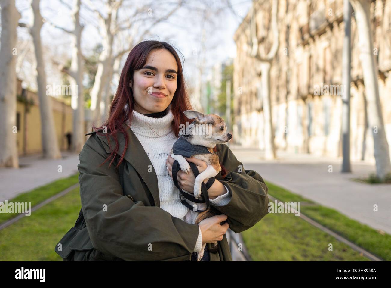 Una donna con lunghi capelli scuri sorride tenendo in mano il suo chihuahua, godendosi una giornata di sole in una strada alberata con un comodo maglione bianco e un jac verde Foto Stock