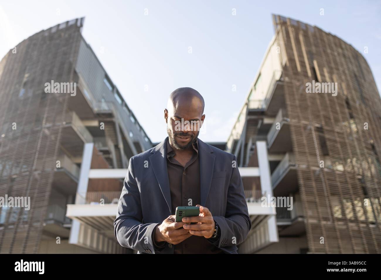 Un uomo cubano d'affari controlla il suo smartphone di fronte a una struttura architettonica contemporanea. È concentrato e vestito con un abito, esemplificando un professionista Foto Stock
