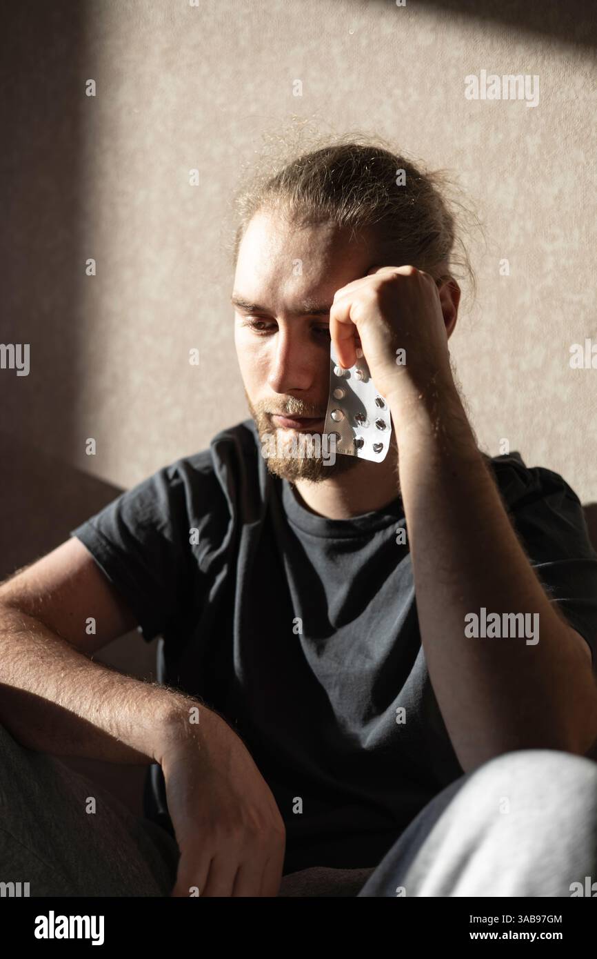 Giovane con la barba che tiene in mano un blister di pillole. Seduto in una stanza, appare profondamente nel pensiero, suggerendo temi di salute o di dicembre Foto Stock
