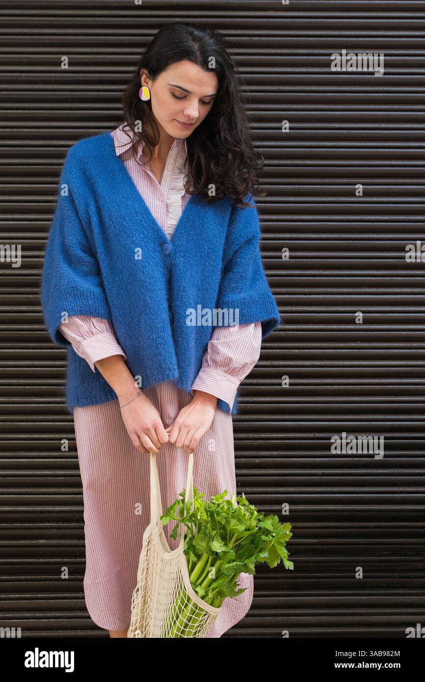 Una donna che indossa un cardigan blu e un abito a righe contiene una borsa in mesh piena di freschi green. I suoi orecchini colorati aggiungono un tocco vibrante e contrastante Foto Stock