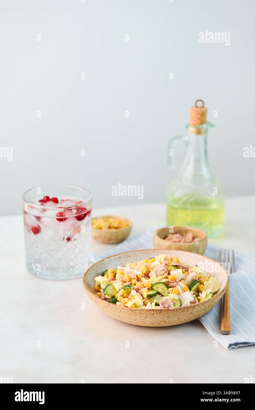 Un'insalata rinfrescante con tonno, mais, cetriolo e guarnire in una ciotola di ceramica, accompagnata da una bevanda con frutti di bosco rossi. Una bottiglia di olio completa il Co Foto Stock
