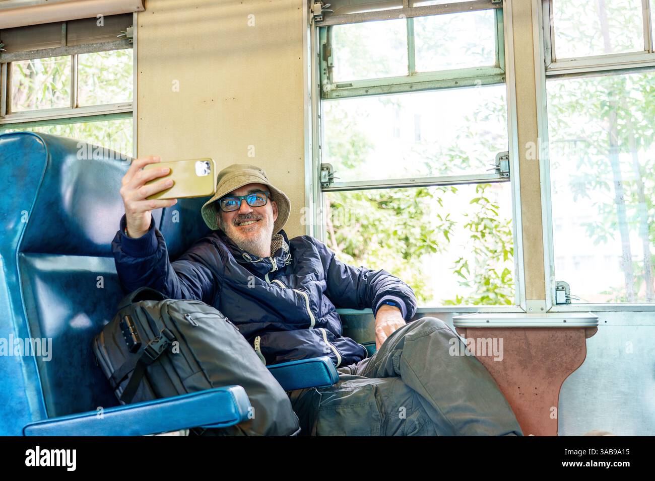 Un uomo allegro e maturo, che indossa un cappello e occhiali, sta scattando un selfie con il suo smartphone mentre è seduto comodamente in un vagone ferroviario. Gli piace la sua jo Foto Stock