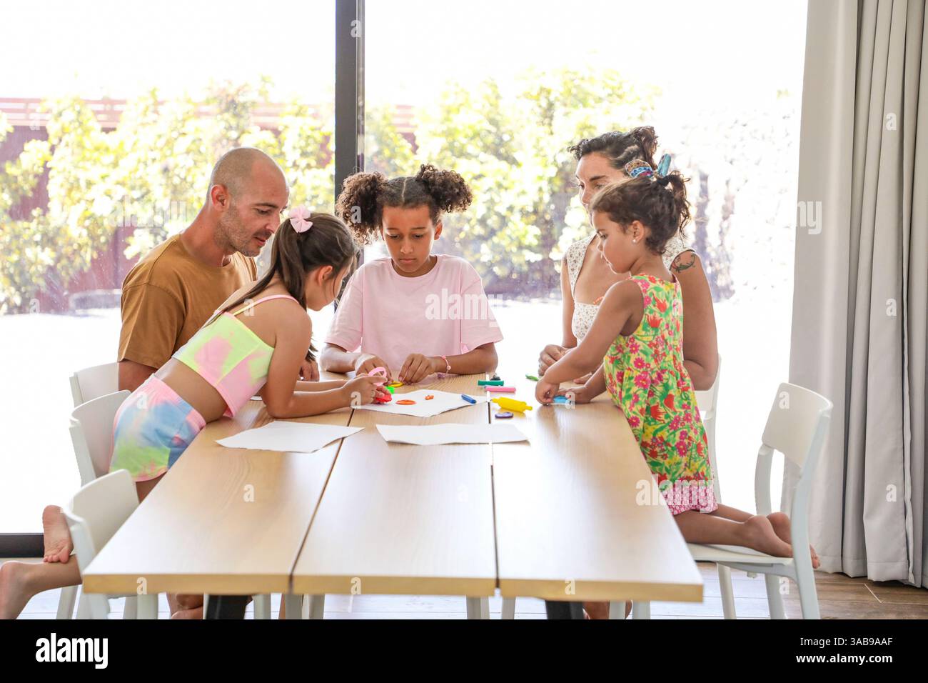 Una famiglia si riunisce intorno a un tavolo di legno, impegnandosi in attività creative insieme. La sala luminosa è piena di luce naturale, che mette in risalto il joyfu Foto Stock