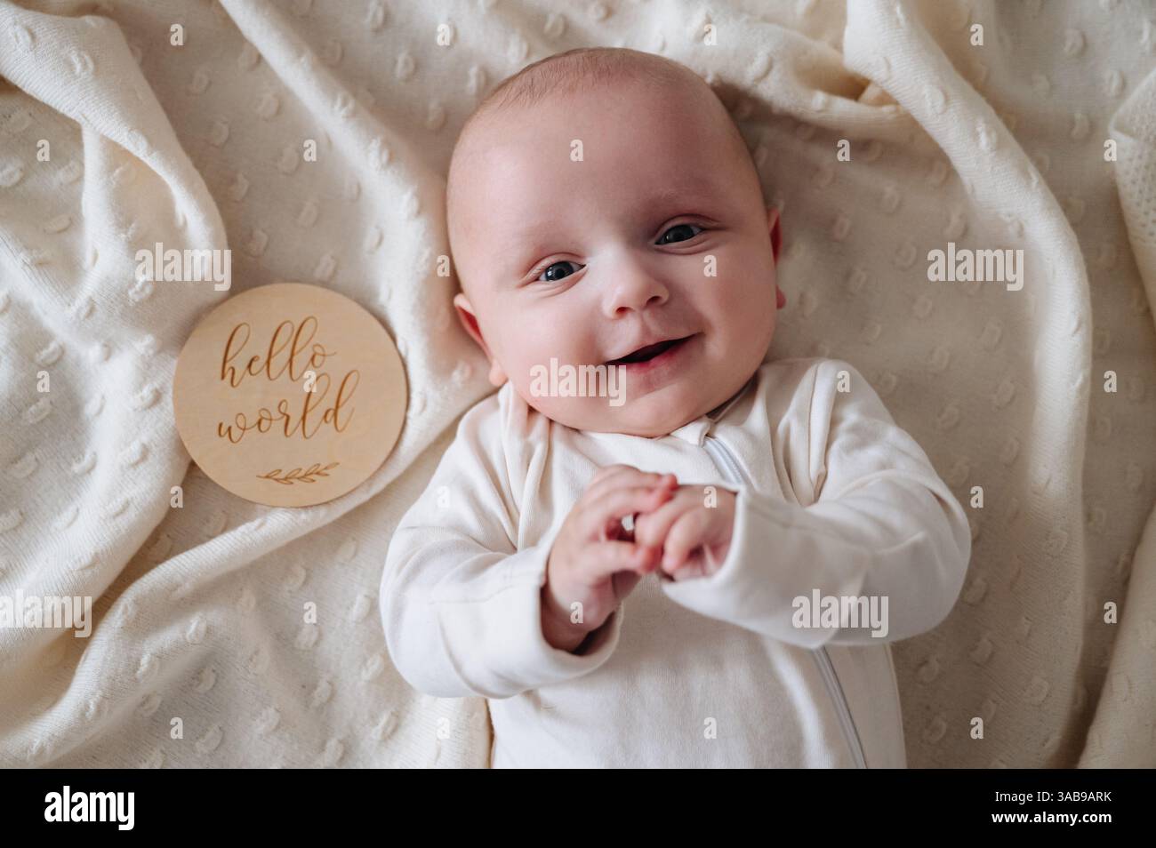 Un bambino sorridente giace su una morbida coperta di crema. Accanto a lui, una targa di legno recita Hello World, catturando il calore e l'innocenza della prima infanzia Foto Stock