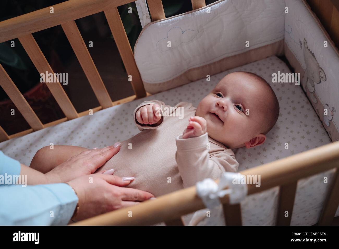 Un bambino carino, con un abito beige, giace in una culla sorridente mentre le mani di sua madre lo toccano delicatamente. L'atmosfera intima enfatizza il calore e la cura. Foto Stock